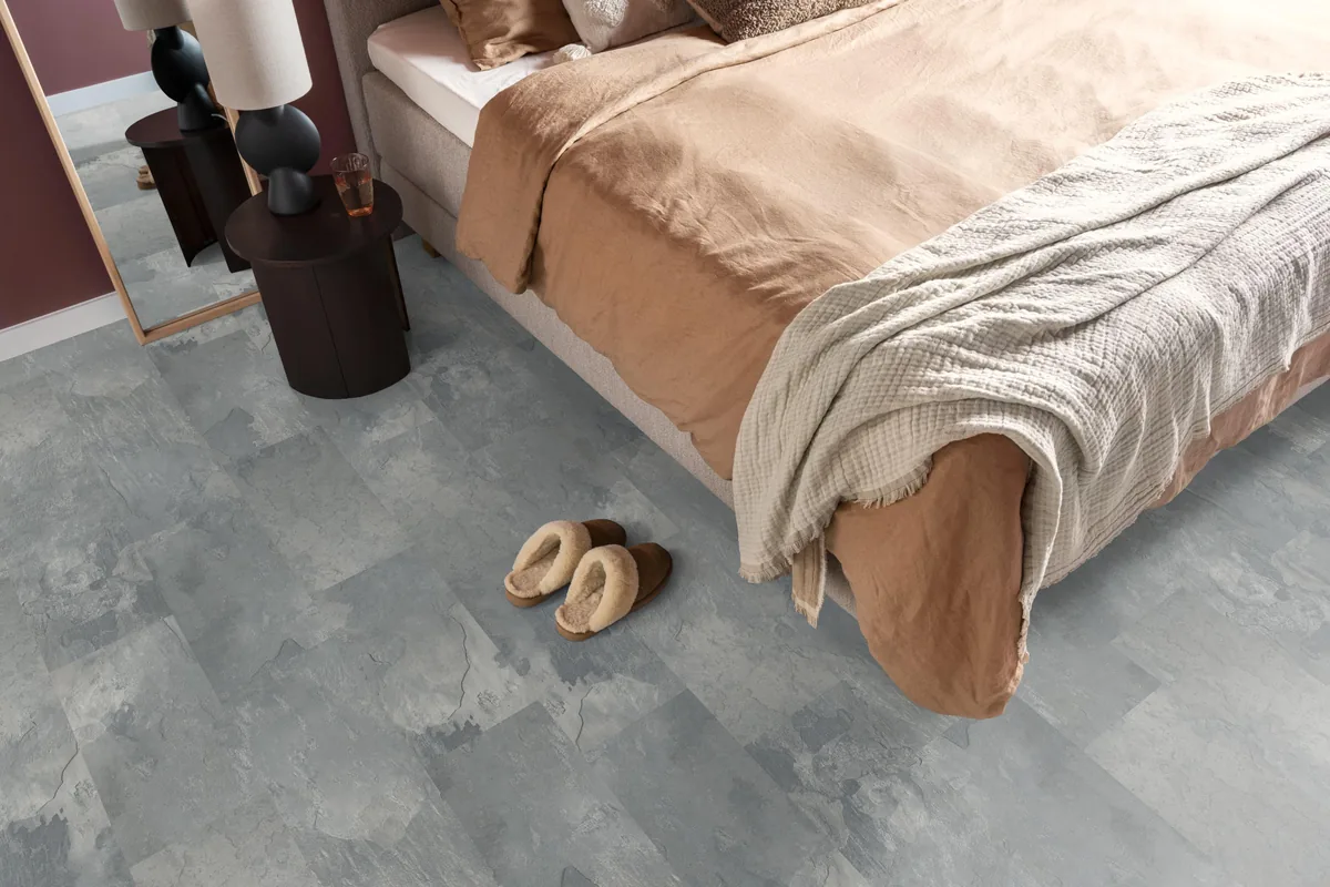 Madison Square Aqua / Expression Aqua middengrijs, tegellaminaat met natuursteenlook in een slaapkamer, met sloffen naast het bed en een donker nachtkastje