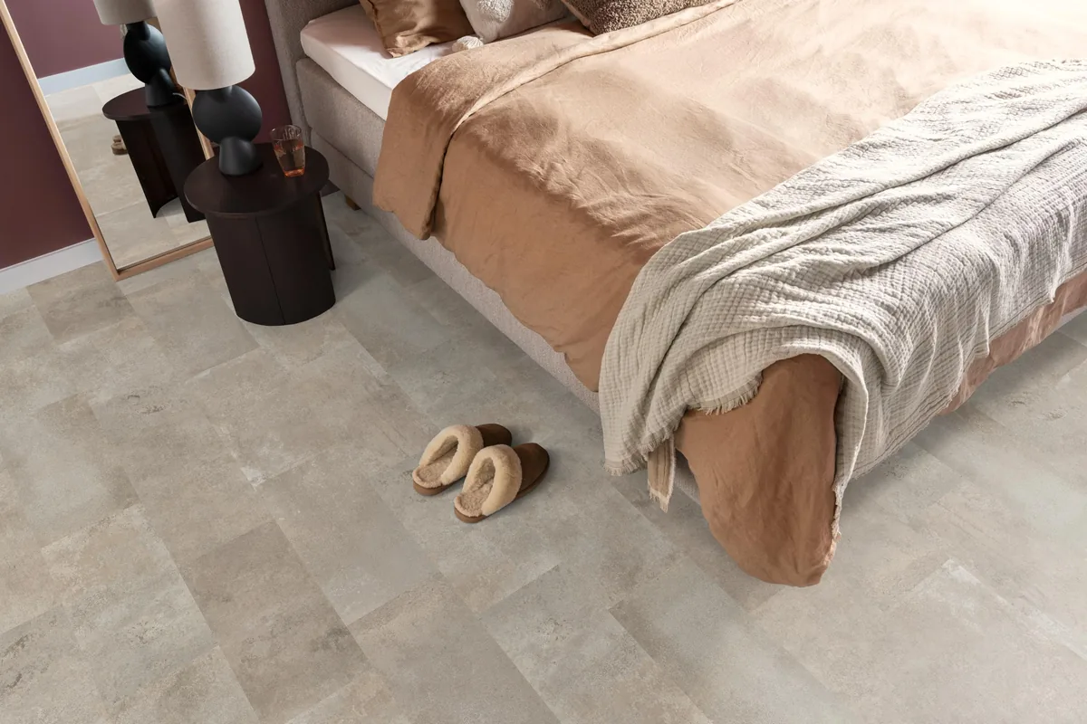 Madison Square Aqua / Expression Aqua zand, tegellaminaat met natuursteenlook in een slaapkamer, met sloffen naast het bed en een donker nachtkastje