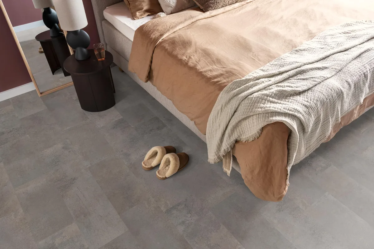 Madison Square Aqua / Expression Aqua grijs, tegellaminaat met natuursteenlook in een slaapkamer, met sloffen naast het bed en een donker nachtkastje
