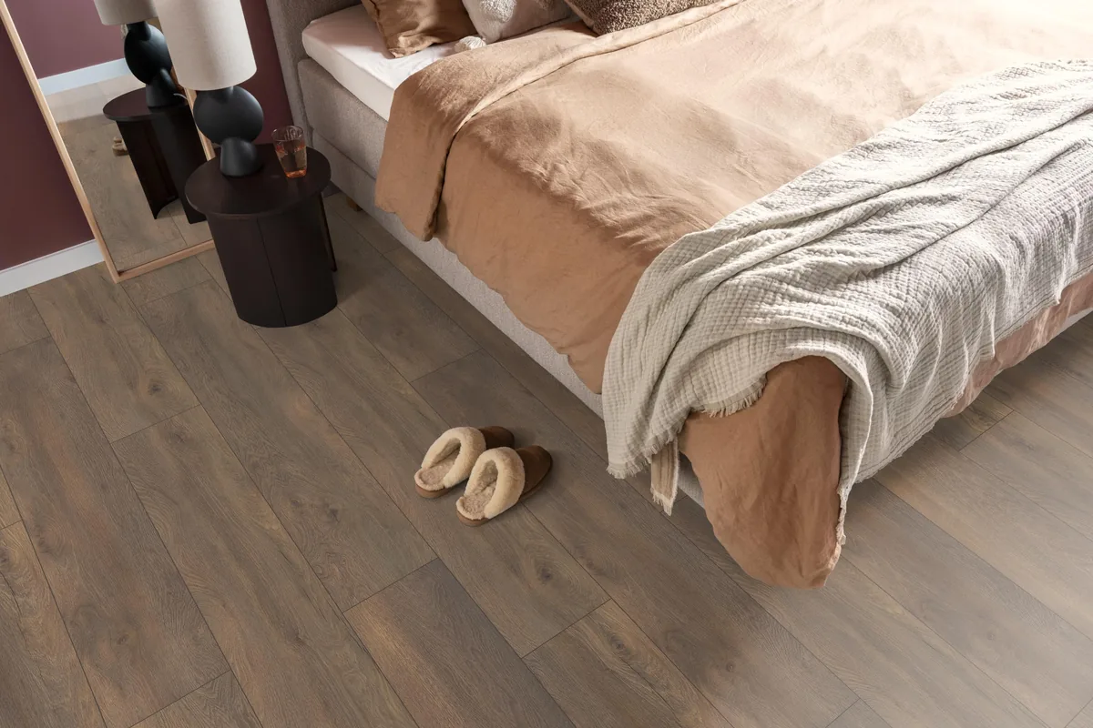 Union Square / Saarland donkerbruin eiken, laminaat in een slaapkamer, met sloffen naast het bed en een donker nachtkastje