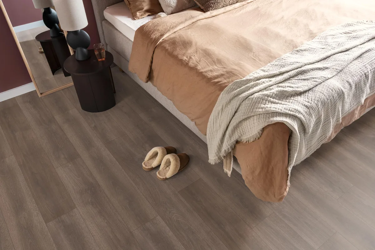 Woodside / Sunny oak donkerbruin eiken, laminaat in een slaapkamer, met sloffen naast het bed en een donker nachtkastje