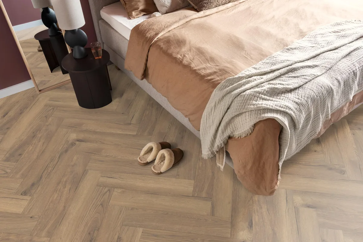 Bayside / Marquant eiken natuur, visgraat laminaat in een slaapkamer, met sloffen naast het bed en een donker nachtkastje