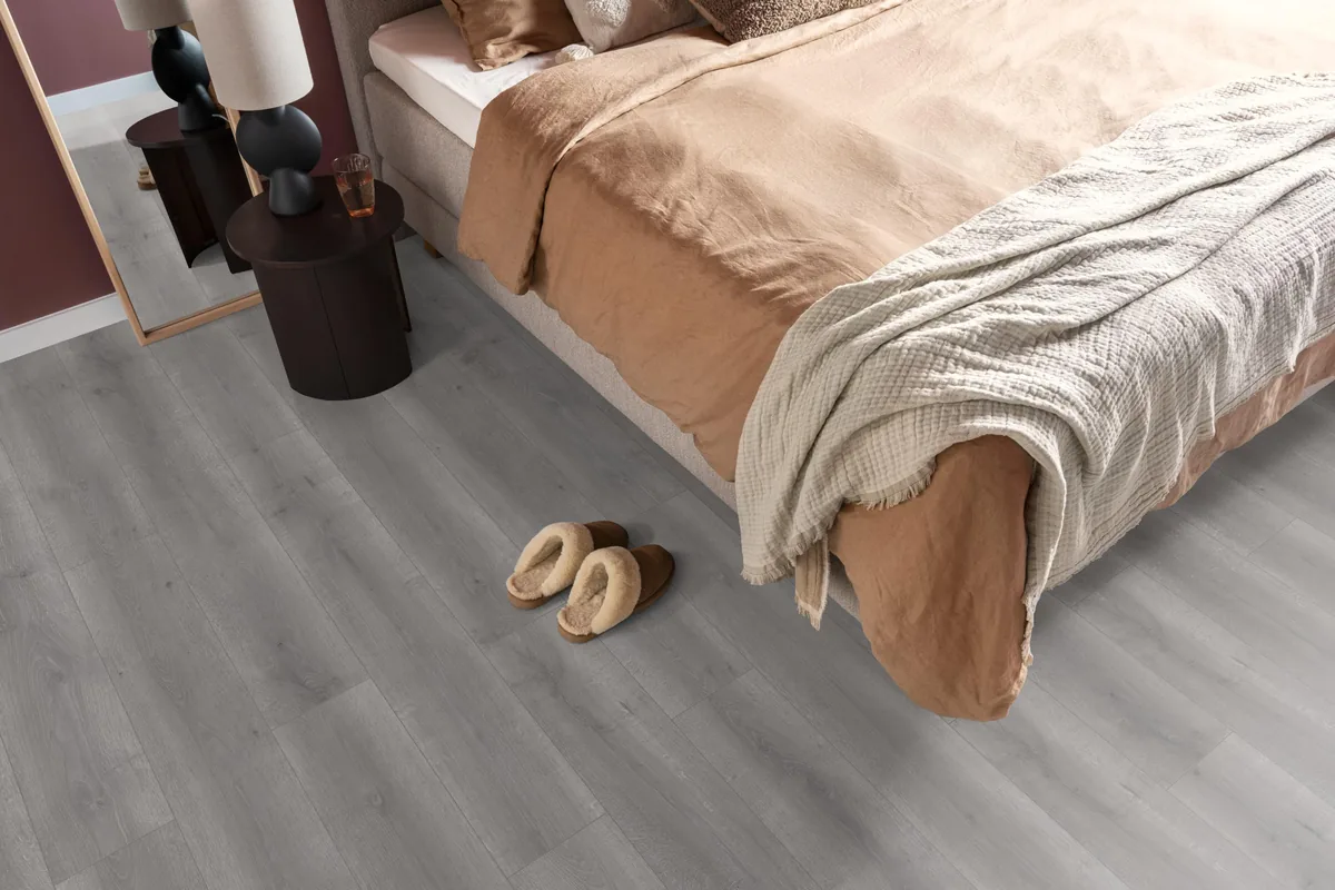 Queens / Elite grijs eiken, laminaat in een slaapkamer, met sloffen naast het bed en een donker nachtkastje