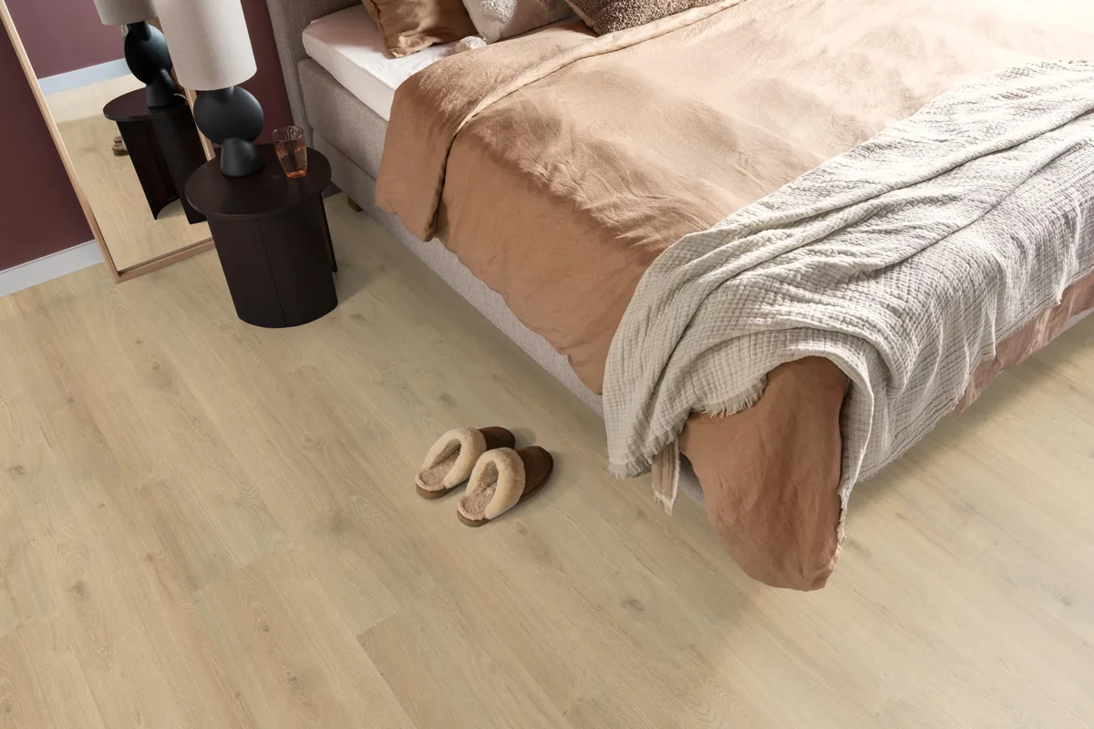 Forest Hills / Aachen beige eiken, laminaat in een slaapkamer, met sloffen naast het bed en een donker nachtkastje