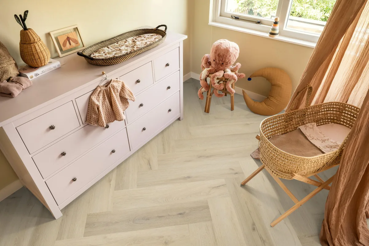 Greenford / Valento Warm beige, visgraat plak pvc in een babykamer, met een witte commode en een rieten wieg