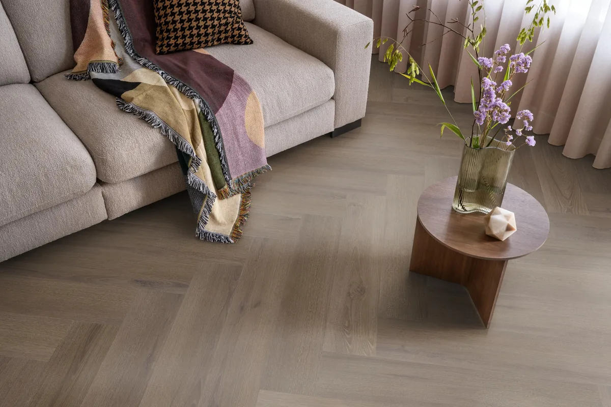 Greenford / Valento Brown, visgraat klik pvc in een woonkamer, met een beige bank en een ronde bijzettafel met bloemen