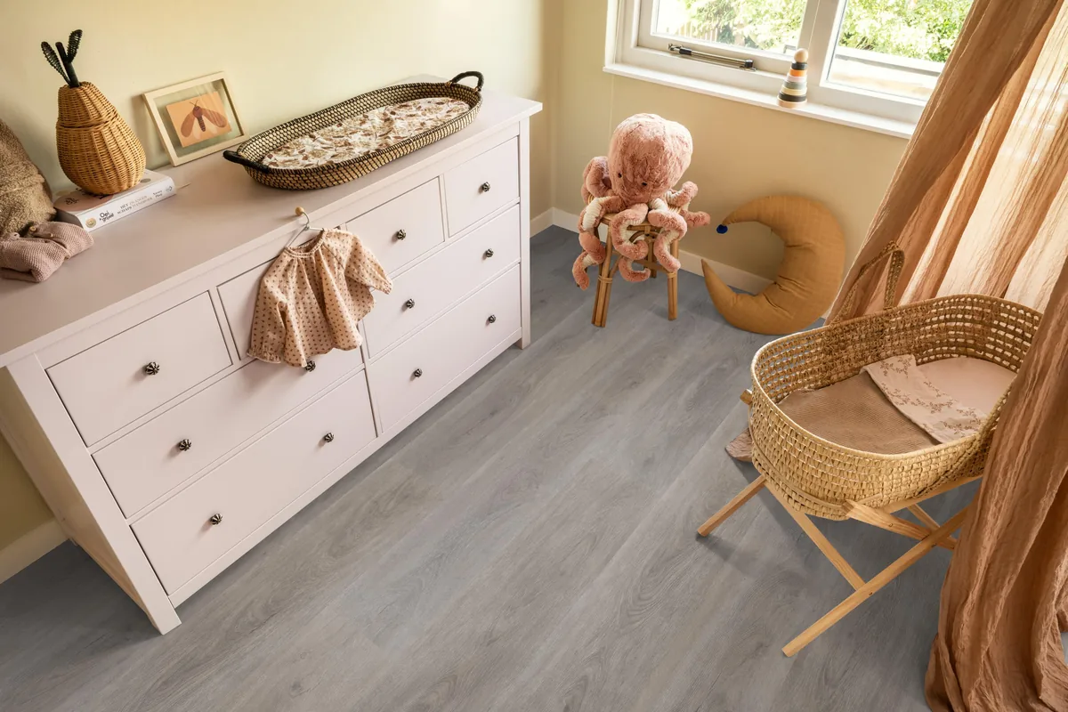 Parramatta / Robusto Grey oak, klik pvc in een babykamer, met een witte commode en een rieten wieg