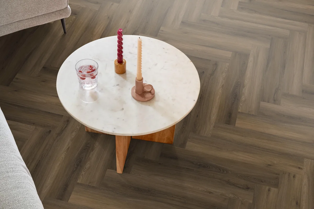 YUP / Spigato Warm brown, visgraat klik pvc in een woonkamer, met een ronde marmeren salontafel en twee kaarsen