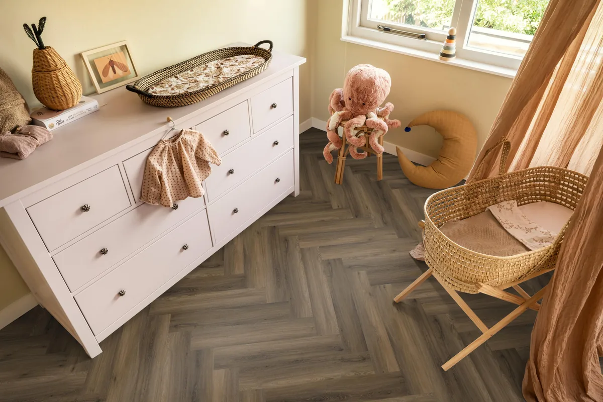 YUP / Spigato Warm brown, visgraat klik pvc in een babykamer, met een witte commode en een rieten wieg
