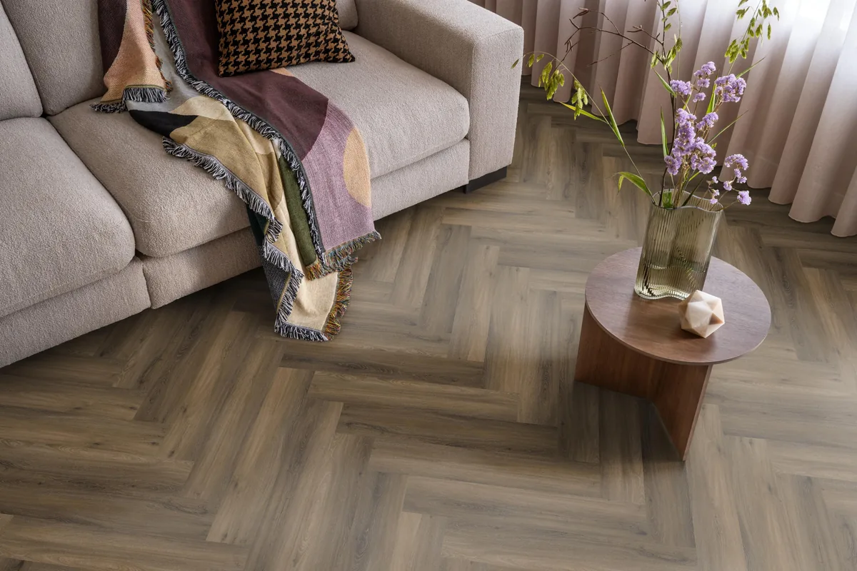 YUP / Spigato Warm brown, visgraat klik pvc in een woonkamer, met een beige bank en een ronde bijzettafel met bloemen
