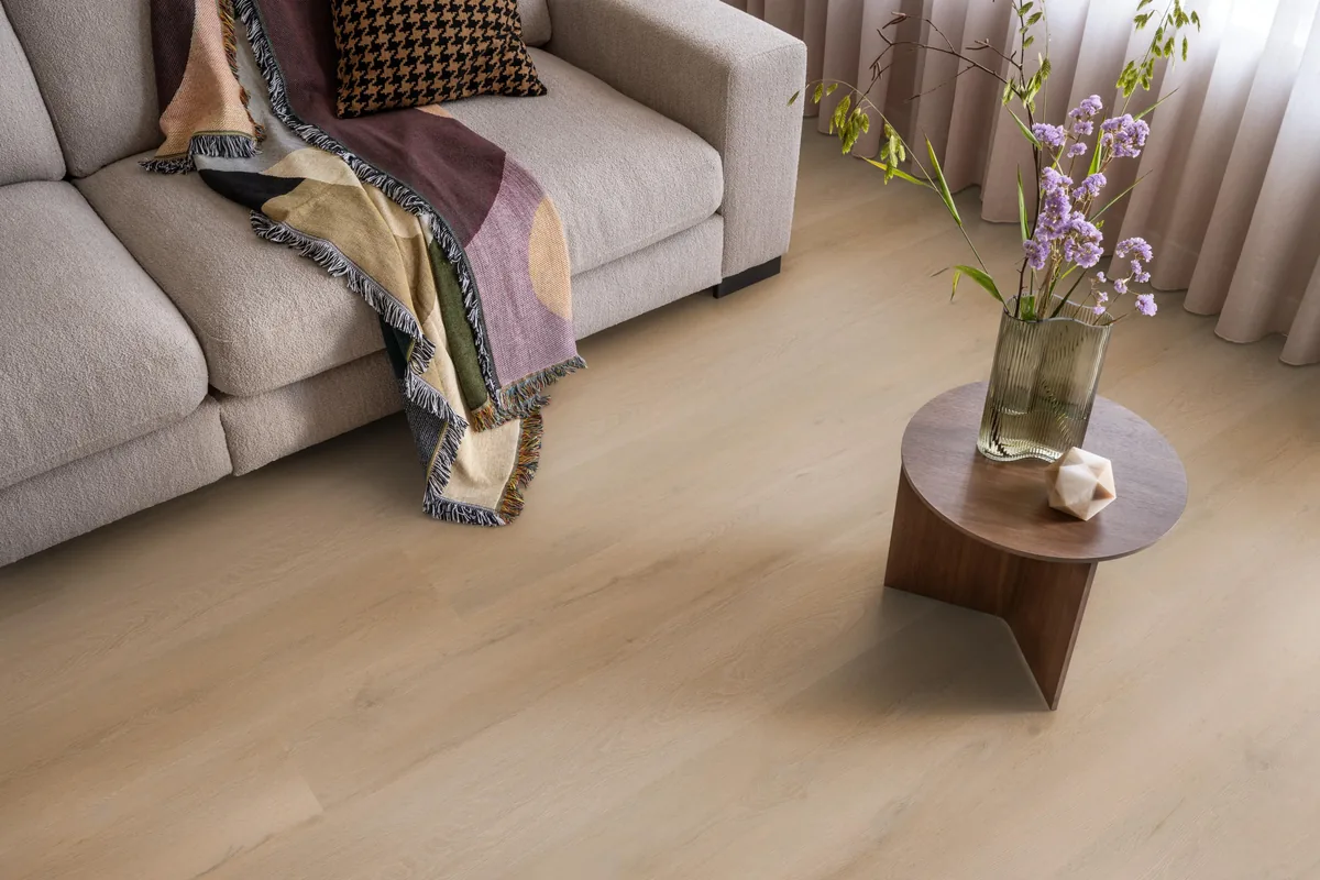 Leyton / Vivero warm oak, klik pvc in een woonkamer, met een beige bank en een ronde bijzettafel met bloemen