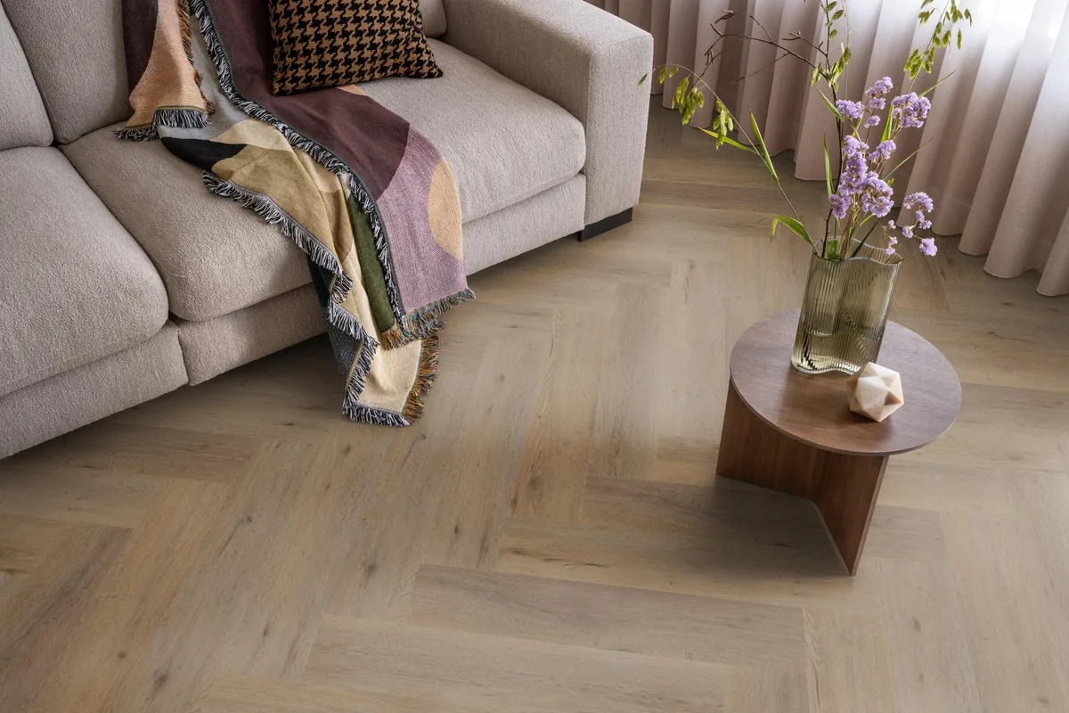 Leyton / Vivero smoky, visgraat plak pvc in een woonkamer, met een beige bank en een ronde bijzettafel met bloemen