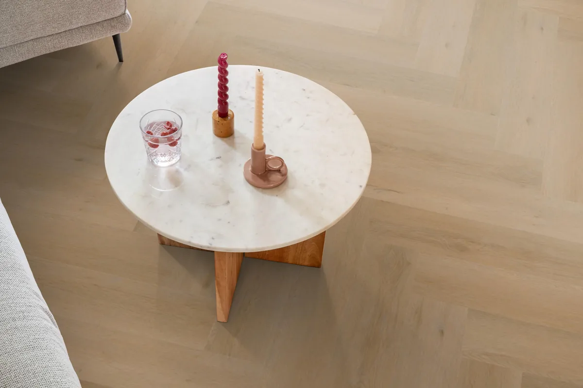 Leyton / Vivero warm oak, visgraat plak pvc in een woonkamer, met een ronde marmeren salontafel en twee kaarsen