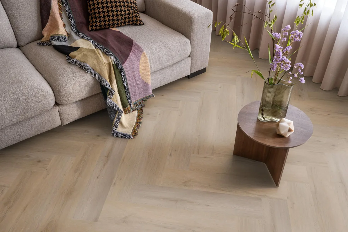 Leyton / Vivero Smoky, visgraat klik pvc in een woonkamer, met een beige bank en een ronde bijzettafel met bloemen