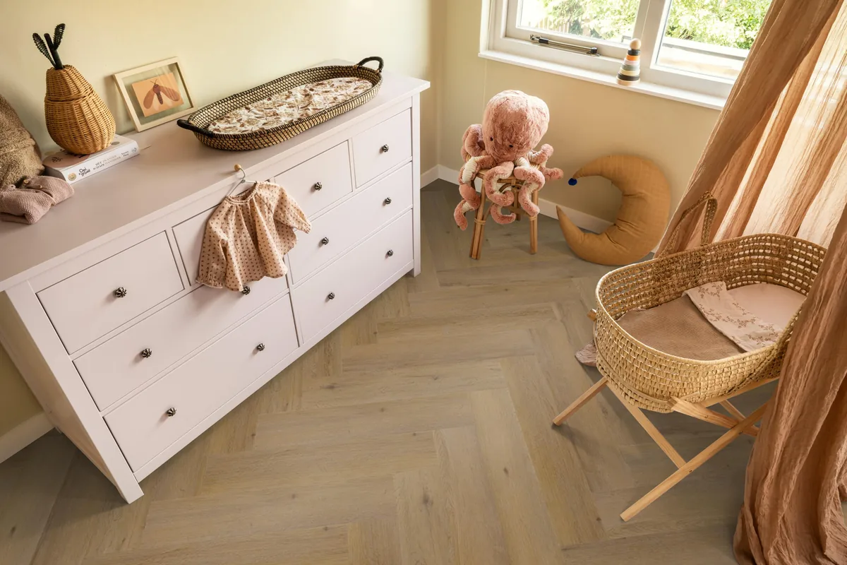 Leyton / Vivero Dark oak, visgraat klik pvc in een babykamer, met een witte commode en een rieten wieg