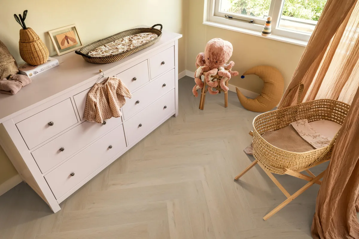 Leyton / Vivero Beige, visgraat klik pvc in een babykamer, met een witte commode en een rieten wieg