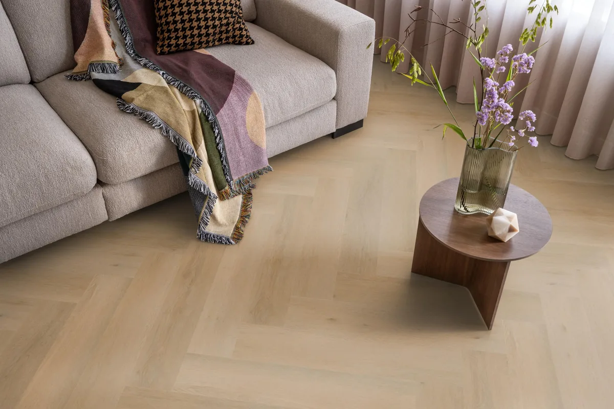 Leyton / Vivero Warm oak, visgraat klik pvc in een woonkamer, met een beige bank en een ronde bijzettafel met bloemen