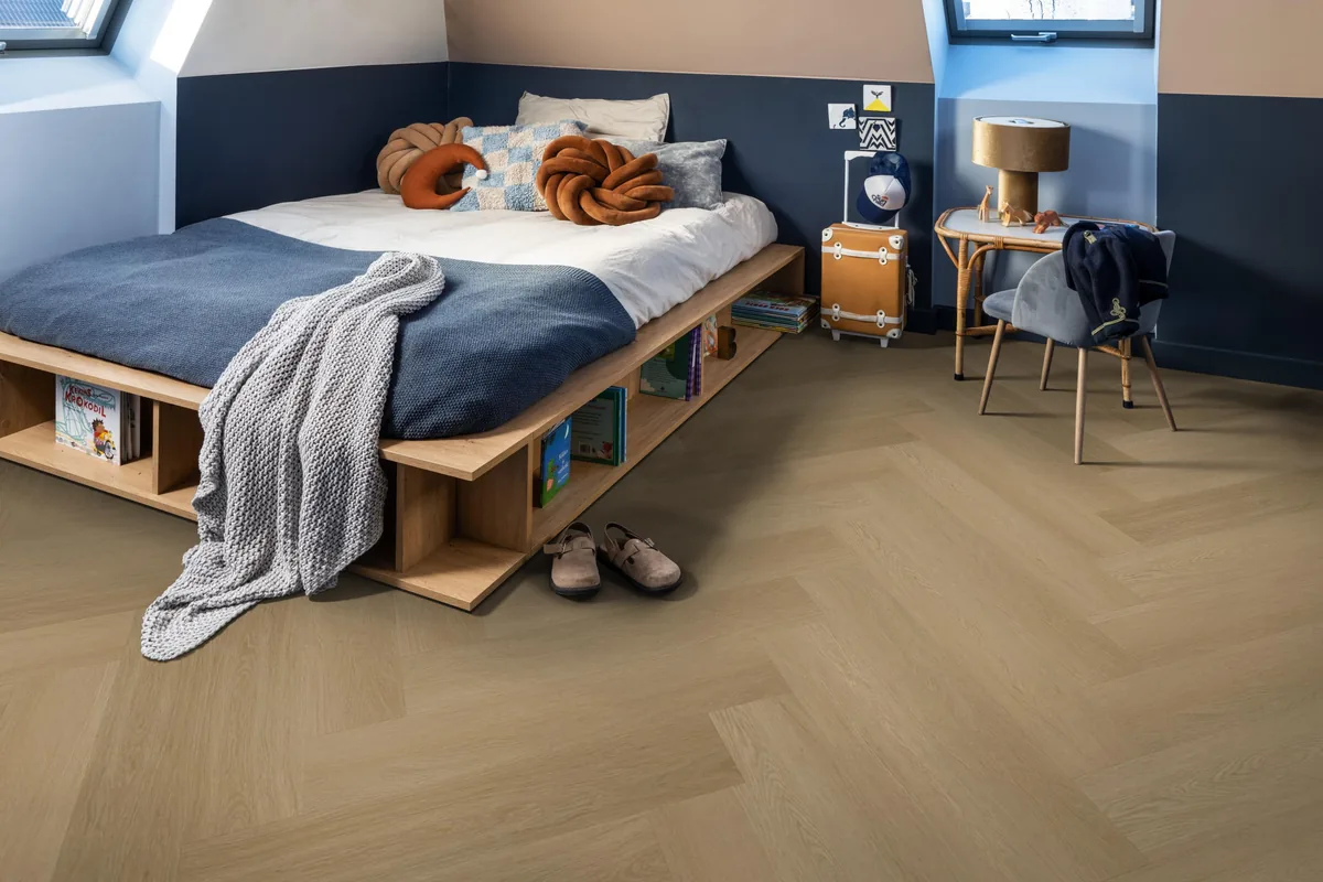 Fulham / Estino warm oak, visgraat plak pvc in een slaapkamer onder een schuin dak, met een houten bedombouw en sloffen ervoor