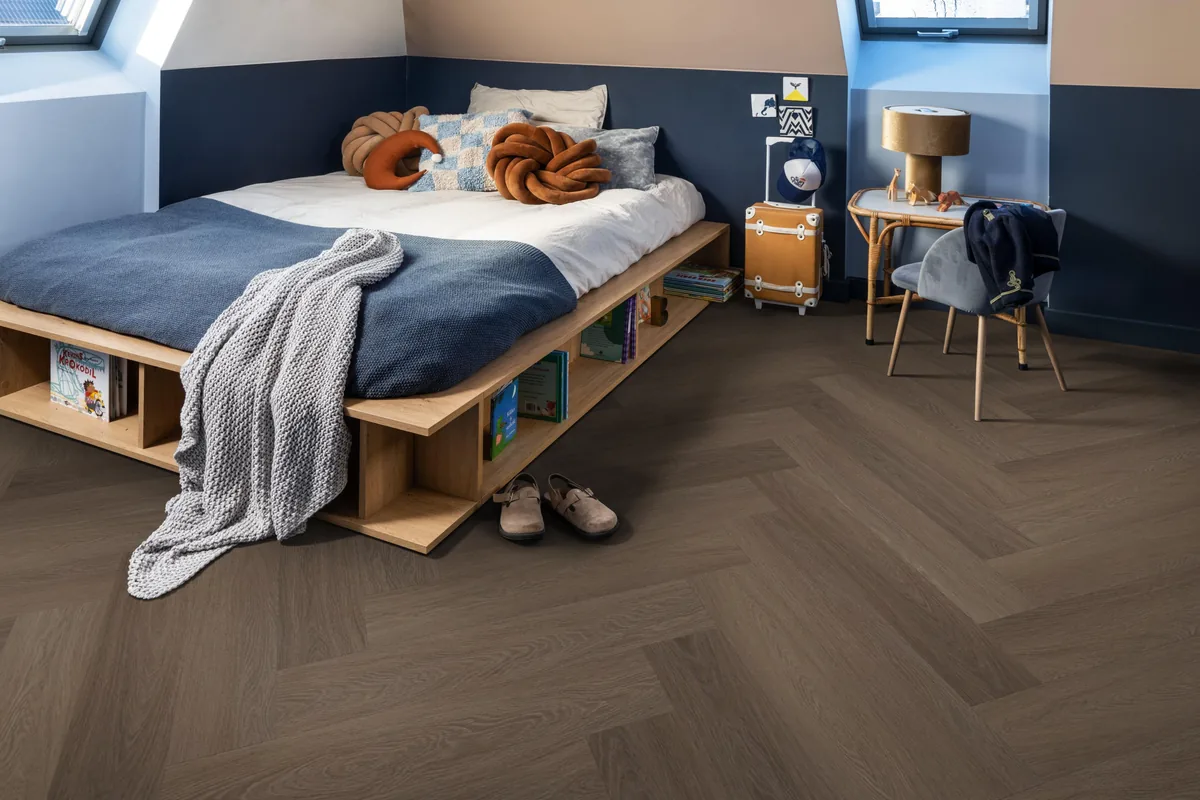 Fulham / Estino brown, visgraat plak pvc in een slaapkamer onder een schuin dak, met een houten bedombouw en sloffen ervoor