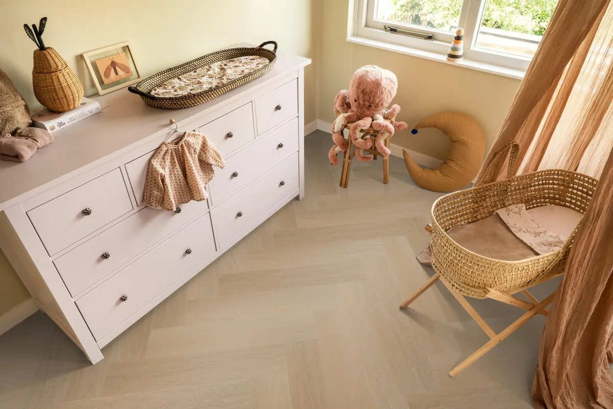 Fulham / Estino Natural oak, visgraat klik pvc in een babykamer, met een witte commode en een rieten wieg