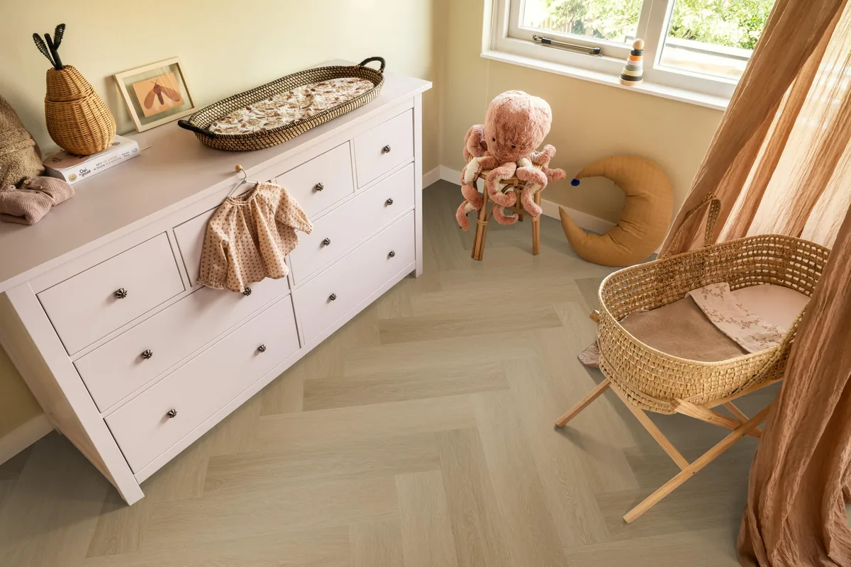 Fulham / Estino Warm oak, visgraat klik pvc in een babykamer, met een witte commode en een rieten wieg