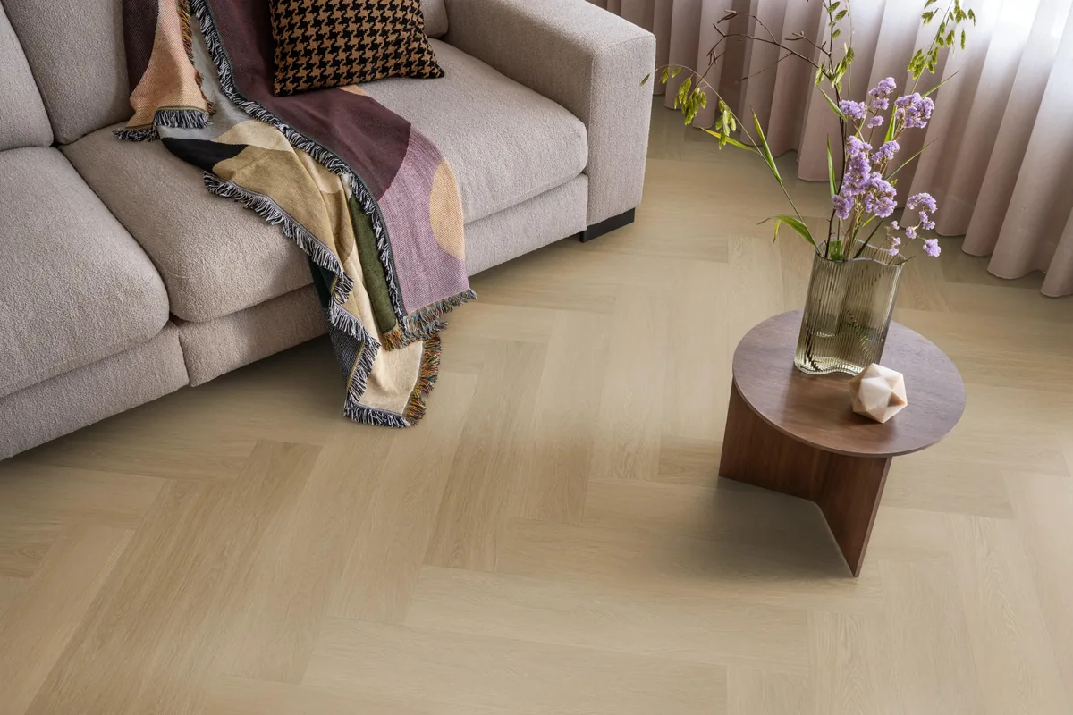 Fulham / Estino Warm oak, visgraat klik pvc in een woonkamer, met een beige bank en een ronde bijzettafel met bloemen
