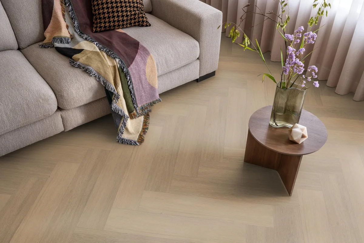 Sutton / Viranto warm natural, visgraat plak pvc in een woonkamer, met een beige bank en een ronde bijzettafel met bloemen