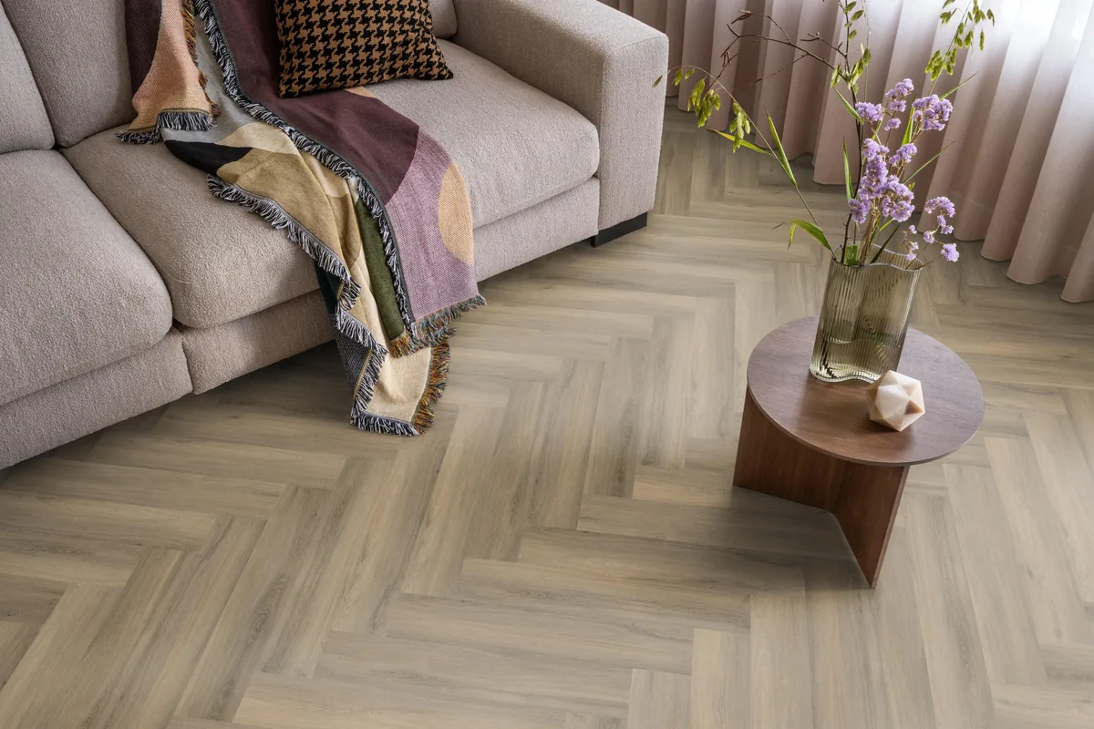 YUP / Spigato light brown, visgraat plak pvc in een woonkamer, met een beige bank en een ronde bijzettafel met bloemen