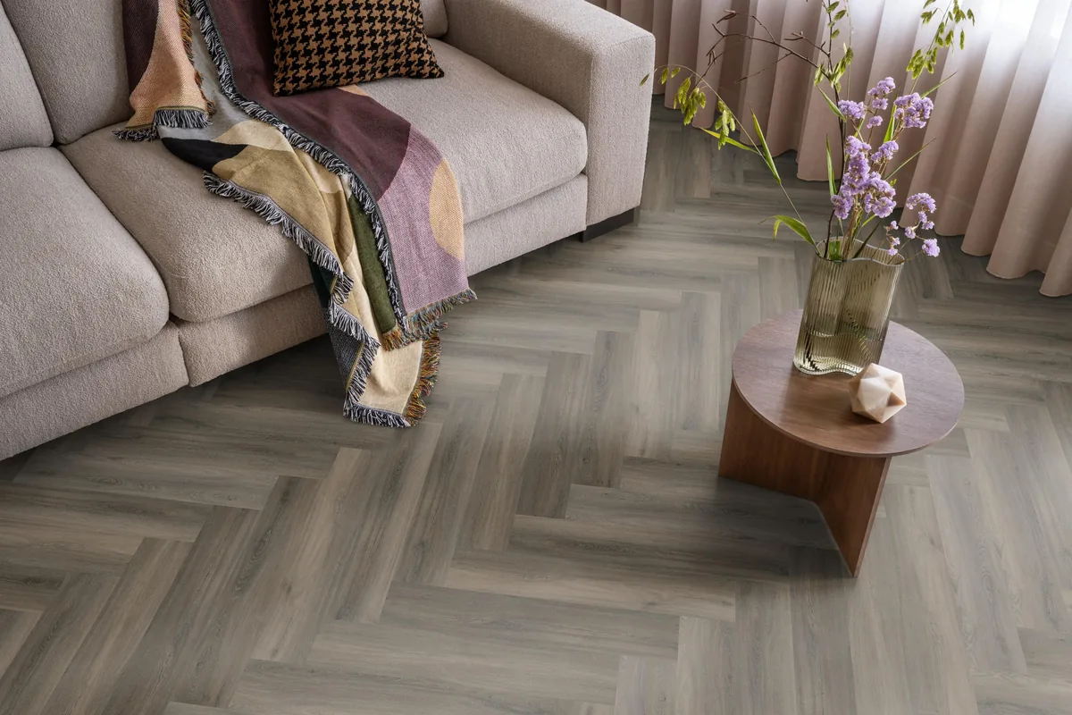 YUP / Spigato dark grey, visgraat plak pvc in een woonkamer, met een beige bank en een ronde bijzettafel met bloemen